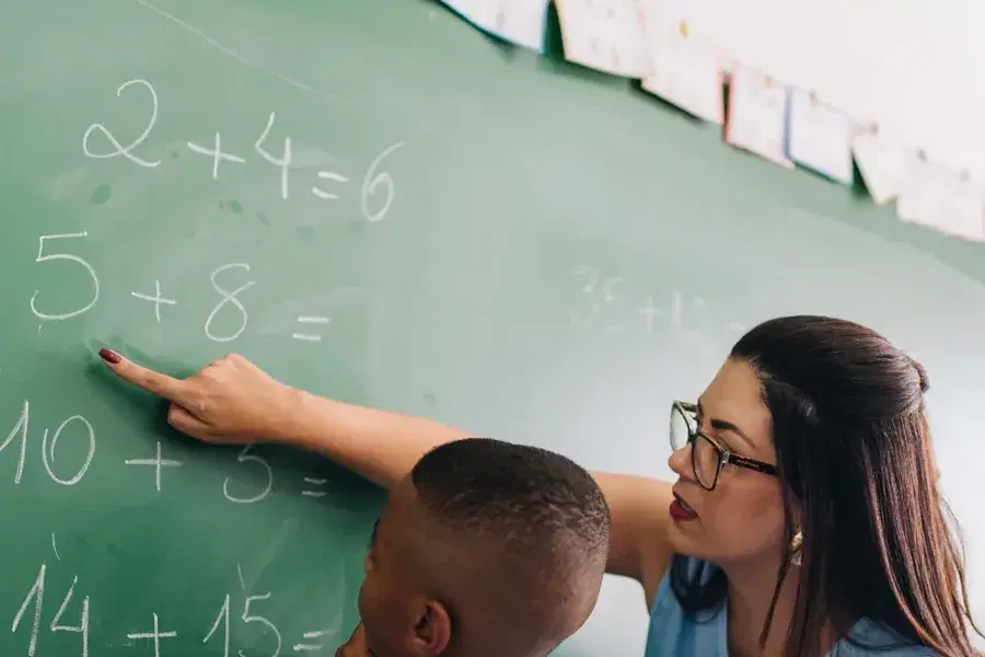 Una profesora señala una cuenta en el pizarrón junto a un estudiante.